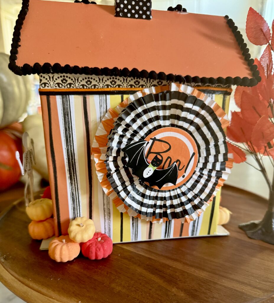 Back of the paper mache Halloween House is covered with striped craft paper and a bunting made from three pleated cupcake paper wrappers, topped with a tiny paper bat.  