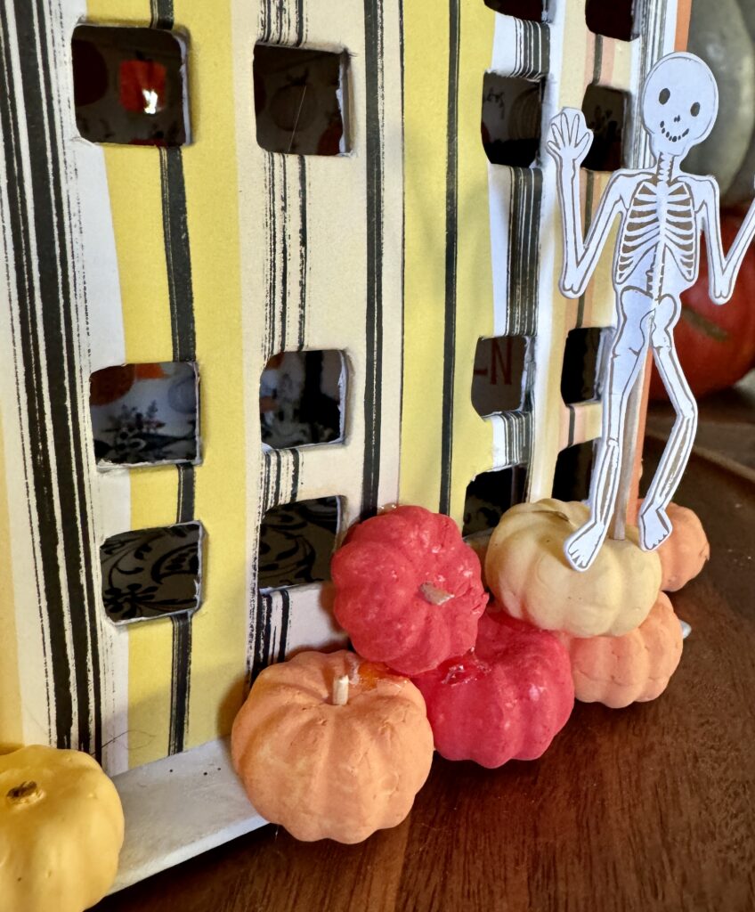 Side View of a Halloween themed paper mache' house. The walls are striped with a yellow, orange, cream and black paper. There are mini-faux pumpkins stacked under the windows. A spooky little skeleton is perched on a stack of pumpkins.  