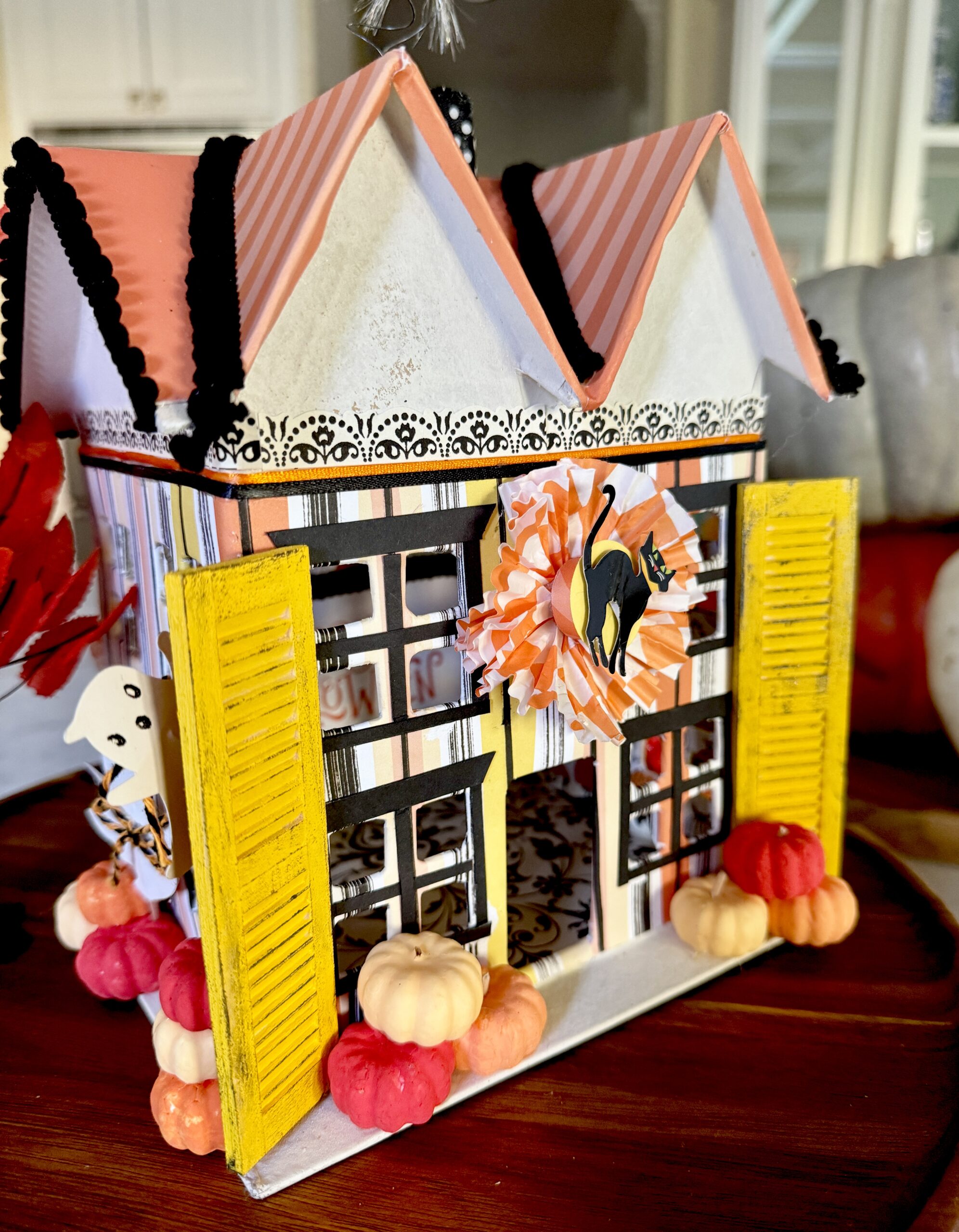 Miniature Halloween House made from a paper mache' base and covered with brightly colored orange, white, yellow and black striped paper. Wooden shutters painted yellow and distressed to look old frame both sides of the house. There's a black cat framed with an orange and white bunting centered above the front door. The roof is a sparkly orange with black fabric trimed edges. Little paper bats are flying out of the black and white polkadot chimney. Tiny pumpkins are stacked around the house.