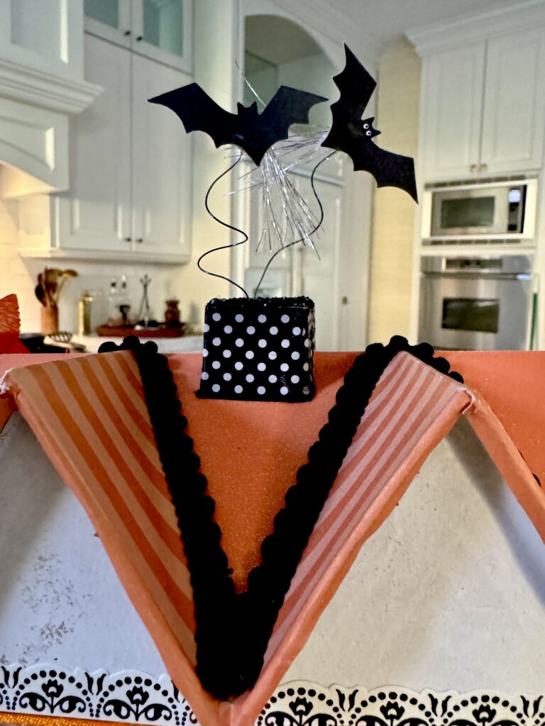 Roofline created in striped and sparkling orange papers and trimmed with black gimp. Bats fly from the black and white ribboned chimney. 