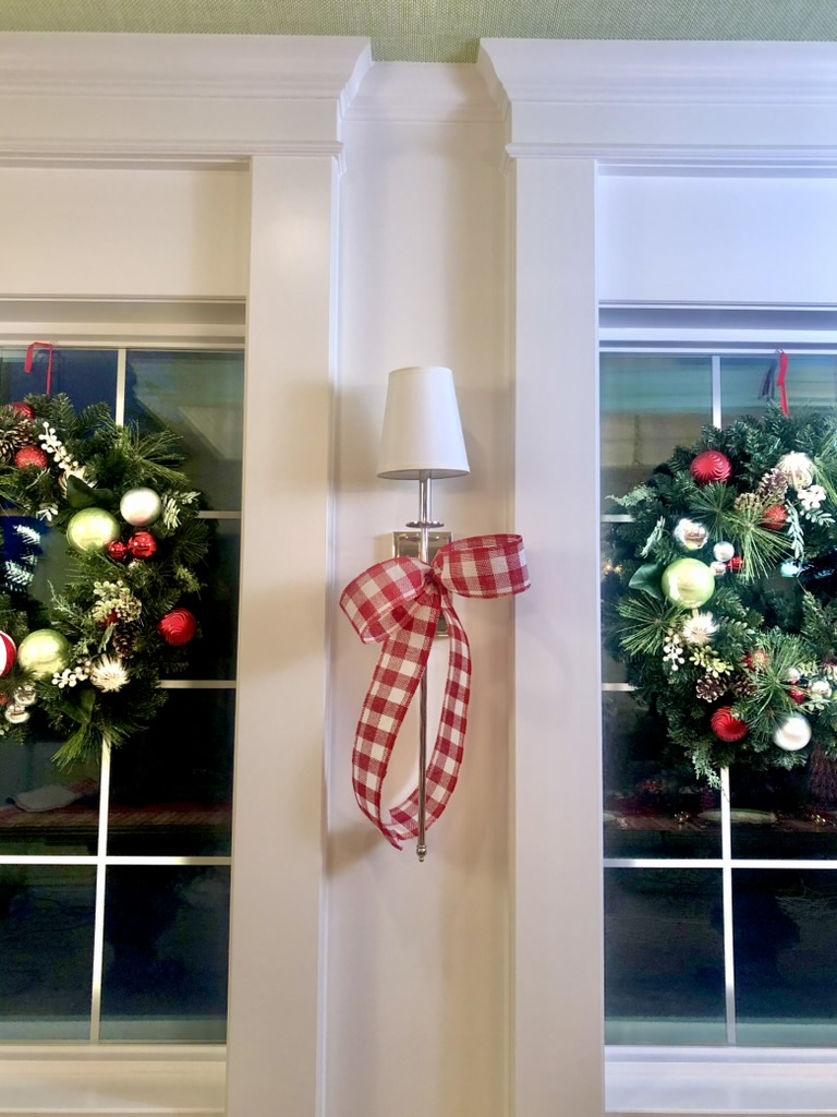 Large Christmas wreaths fill two, side by side windows with a long silver light sconces centered between them. The wall sconce is draped in a large red & white buffalo checked bow. 