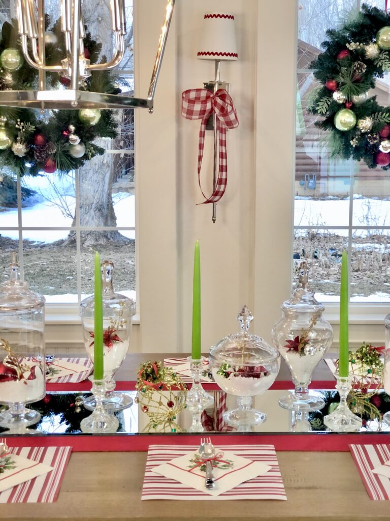 A dining room and it's table all decorated for Christmas with shiny glass chandles sticks, appothacary jars filled with sparkly Christmas ornaments that are all set uptop a glittering mirrored table runner. The room is lit with long silver candle sconces draped in large red & white checked bows. 