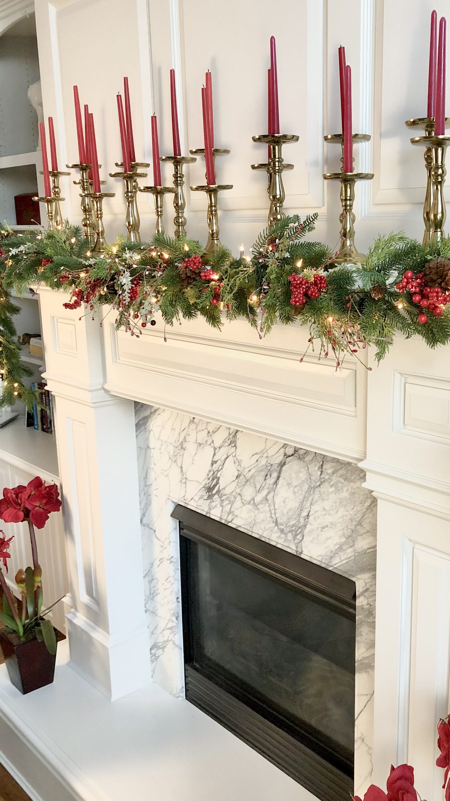 Living Room mantel decorated with christmas garland of cedar with red berries. Dozens of red taper candles in large brass candle sticks are placed ontop the mantles wide shelf.