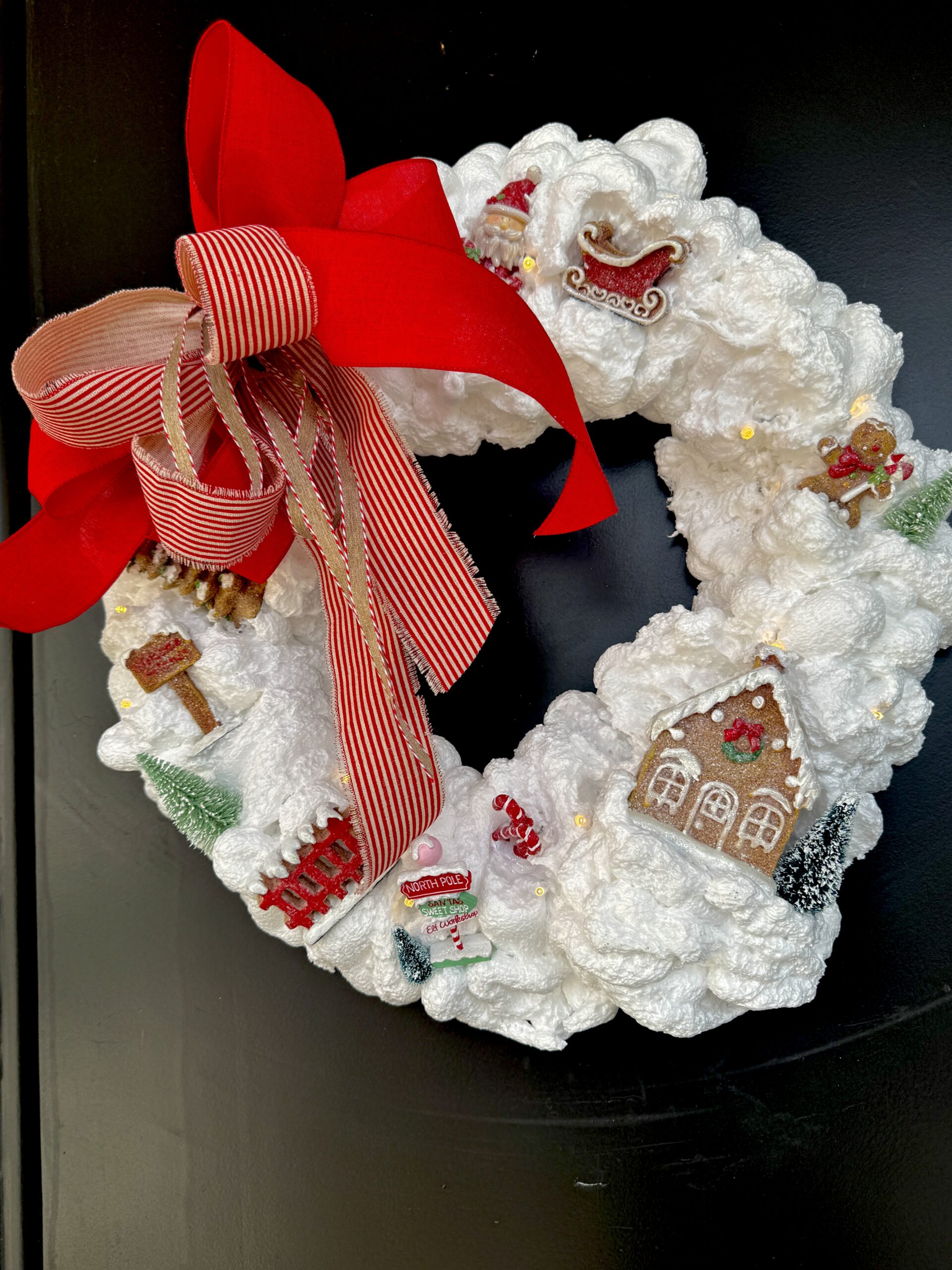 Holiday wreath made from spray foam. Decorated with gingerbread houses, a gingerbread Santa with sleigh and gingerbread people. A festive red bow is tied at the top.