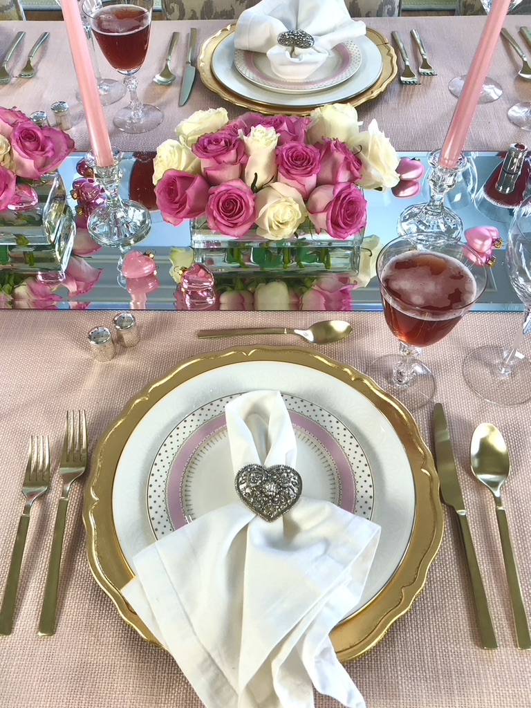 Valentines Day table setting. A soft pink table cloth with a mirroed table runner set the stage for layers of gold and pink china and utensils. Mounds of pink and white roses are punctuated with light pink taper candles in low glass containers.