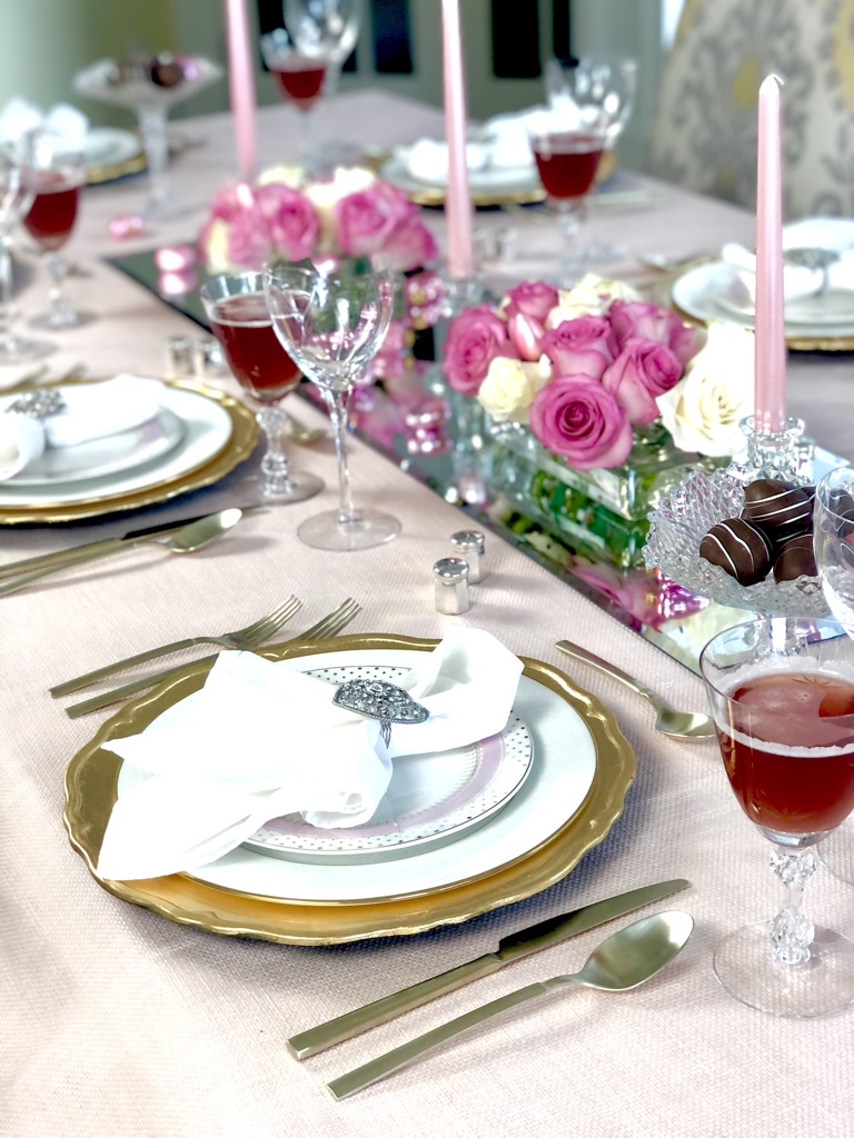 Valentines Day table setting. A soft pink table cloth with a mirroed table runner set the stage for layers of gold and pink china and utensils. Mounds of pink and white roses are punctuated with light pink taper candles in low glass containers.