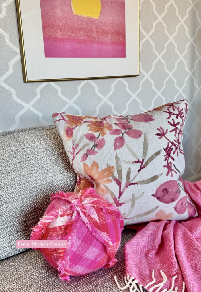 Ball Pillow made in a sweater knit pink & orange buffalo plaid. Sitting on the sofa of a lovely living room with a printed grass cloth wallpaper.