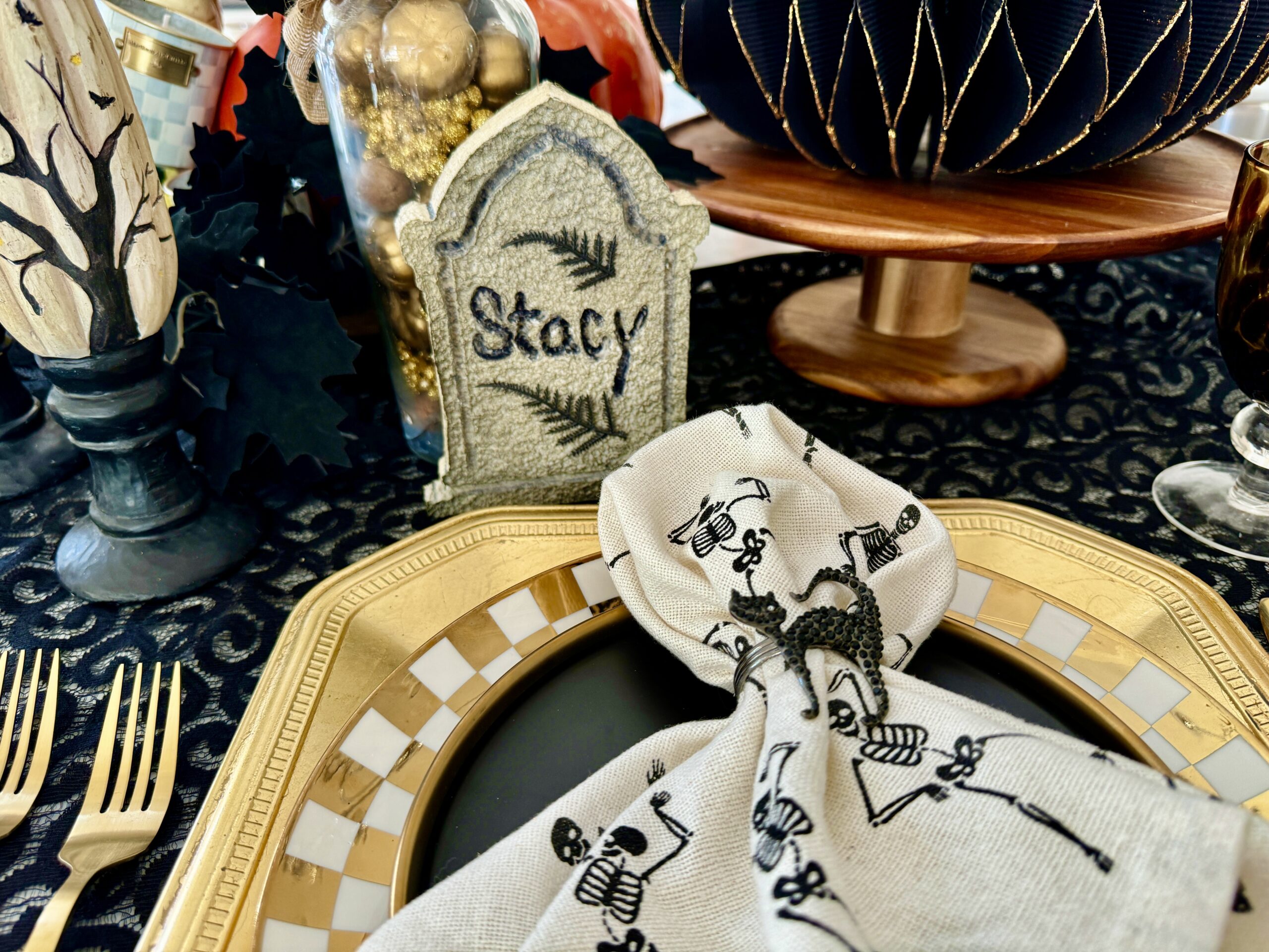 A Halloween table set with gold and black dinnerwear. There is a cream linen napkin with skeletons and a glittery black cat napkin ring. The place cards are realistic mini headstones made from foam.